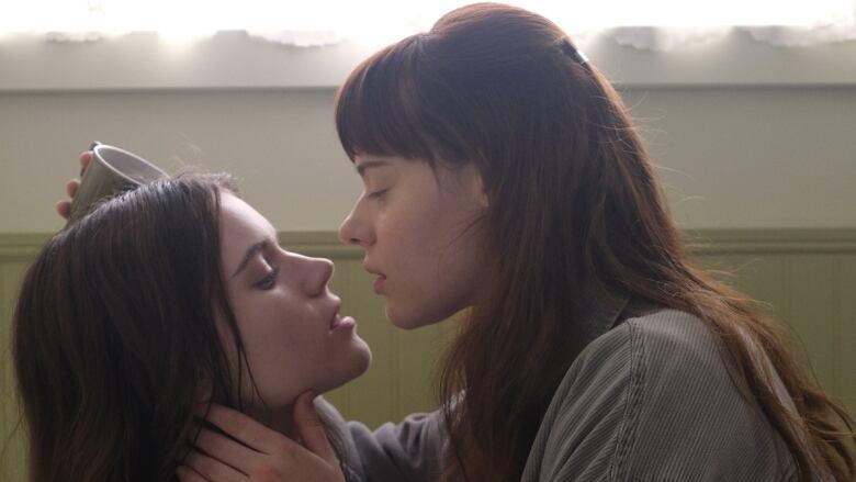 Two women sit in a bathtub, posed as if they are about to kiss. 