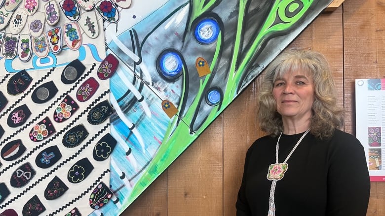 A woman stands against a wooden wall. At her side hangs an art piece. She is dressed in black with and has a determined expression.