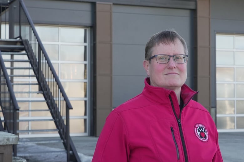 A woman with glasses and in a red jacket stands in front of a building.