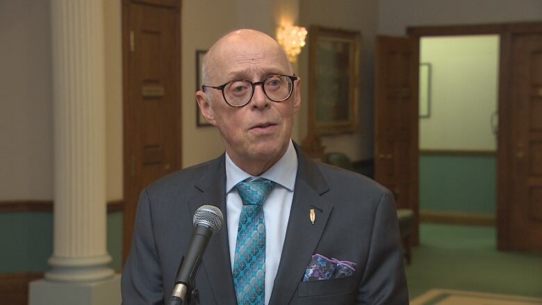 A man with glasses wearing a suit faces the camera, looking to the right of the frame.