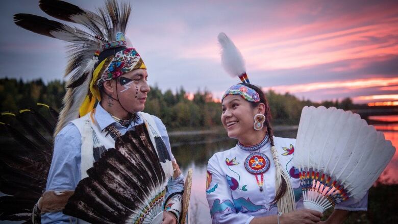 A Cree man and a woman stand smiling at each other, dressed in full-regalia