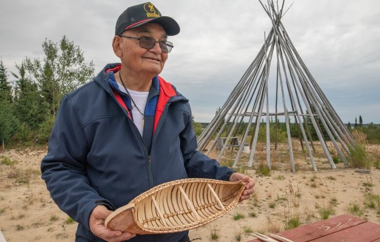 A Cree elder holds a small wooden canoe in front of the a teepee frame.