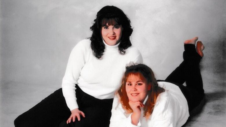 A portrait of two young women, one with dark hair and one with blonde hair, lounging in a studio with white shirts and dark pants  