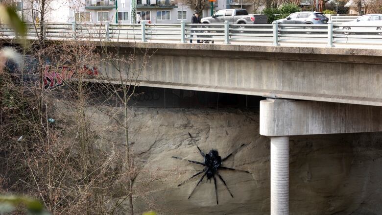 A large black spider sculture, about a few meters in length, is stuck to a rock formation underneath an overpass. 