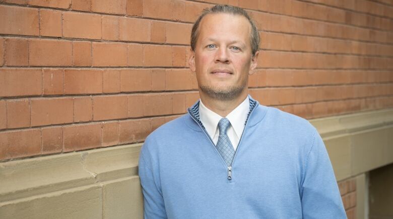 A man wearing a sweater stands in front of a brick wall.