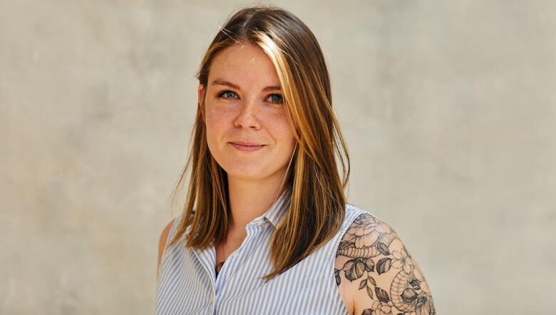 A young woman smiles in this headshot. She has brown hair with a blonde streak. She is wearing a button up shirt that has stripes. She has floral tattoos on her arm. 