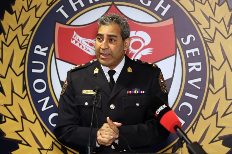 A man in a police uniform stands before a podium speaking with his hands clasped together. Behind him on a screen are the words 'Honour through service.' 