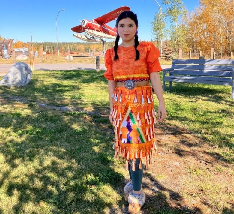 A young girl with braided hair stands outside wearing a bright oranage and yellow dress adorned with jingle cones.