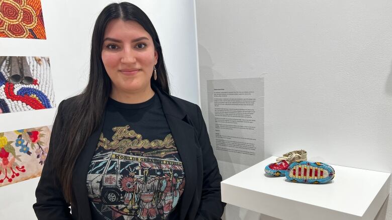 A woman smiles to the camera next to a pair of quilled, baby moccasins.