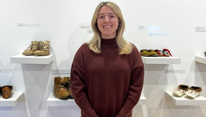 A woman smiles to the camera. Behind her, there are several pairs of moccasins displayed.