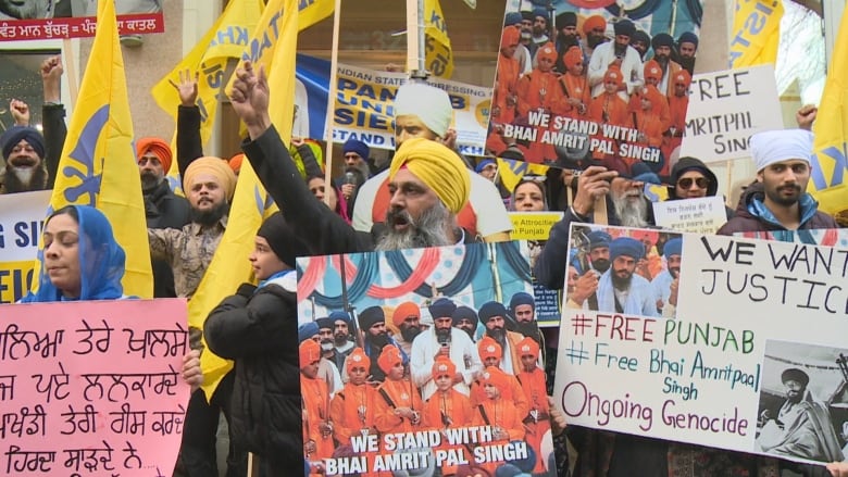 A crowd of Sikh people hold placards and yellow and blue flags with their arms in the air