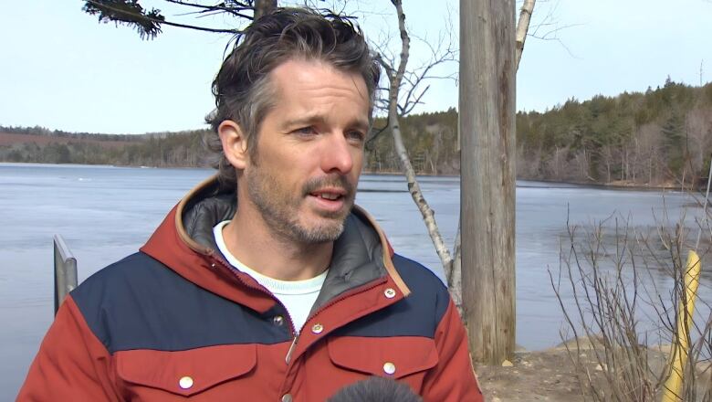 A man with medium-length grey hair and facial stubble stands on a lakeshore in a red jacket