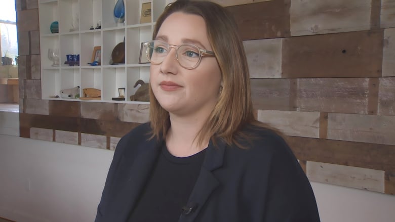 A white woman with shoulder-length light brown hair and glasses stands in front of a wood-panelled wall with shelving.