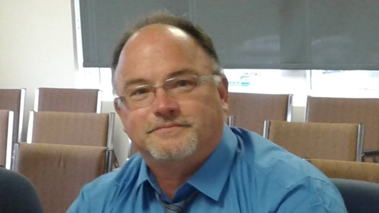 A man in a blue shirt and striped blue tie signing papers at a table. He's balding and wearing glasses.