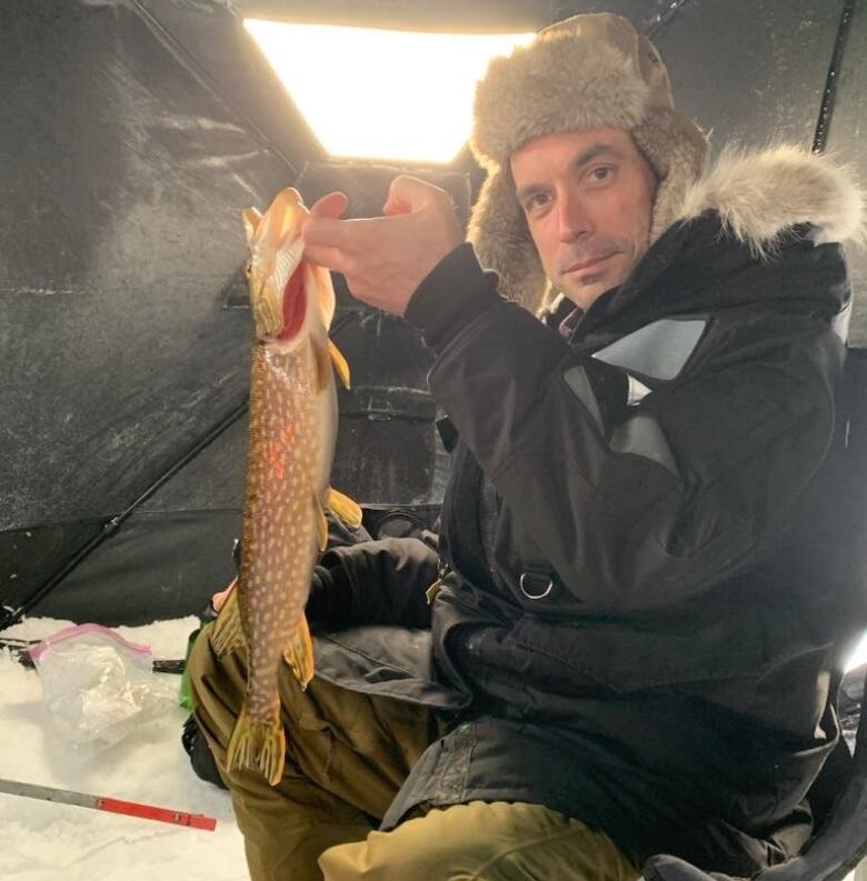 A man in a parka and hat sits inside an ice-fishing hut holding up a fish.