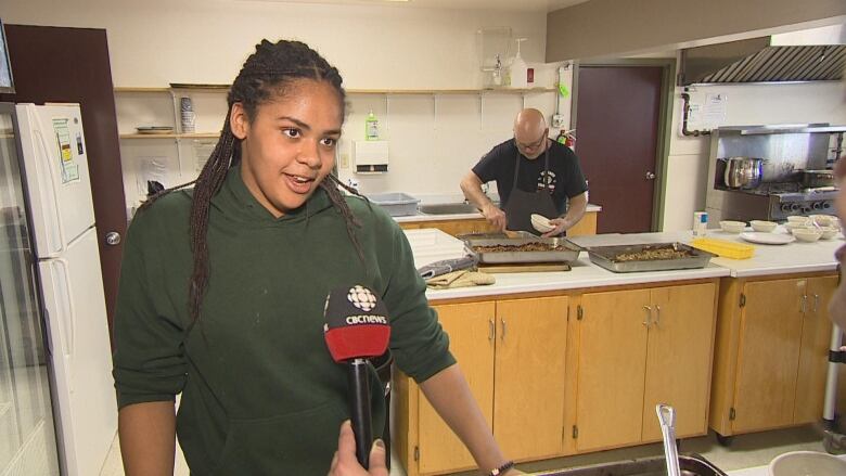 A young woman in a green sweater stands in a kitchen.