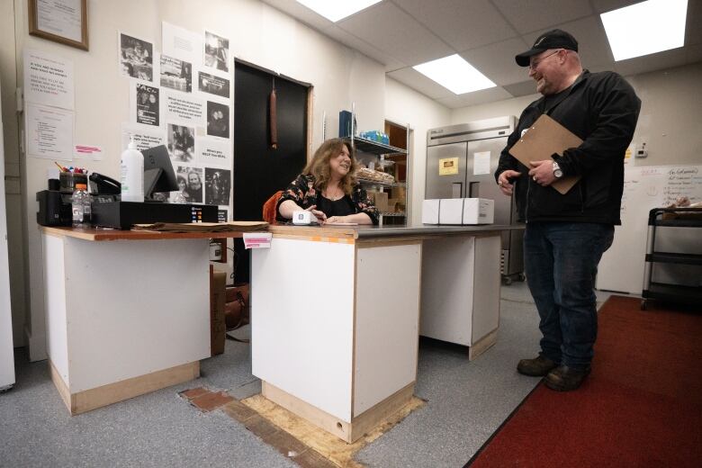 Brigida Crosbie sits at her desk in the office of Tydel Foods as she smiles while greeting a man with a clipboard. 