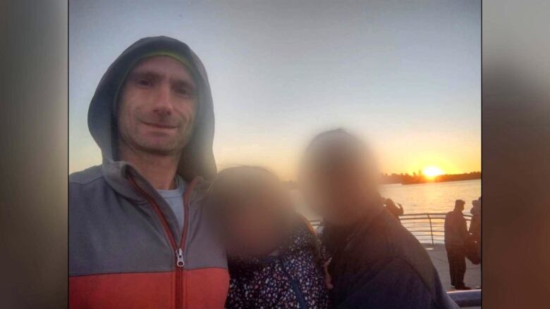 A smiling man poses for a photo in front of a sunset with a woman and girl whose faces have been blurred for privacy.