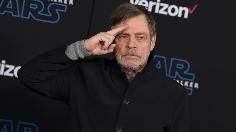 A man in a black, high-collared, button down shirt raises his right hand to his forehead in a salute.