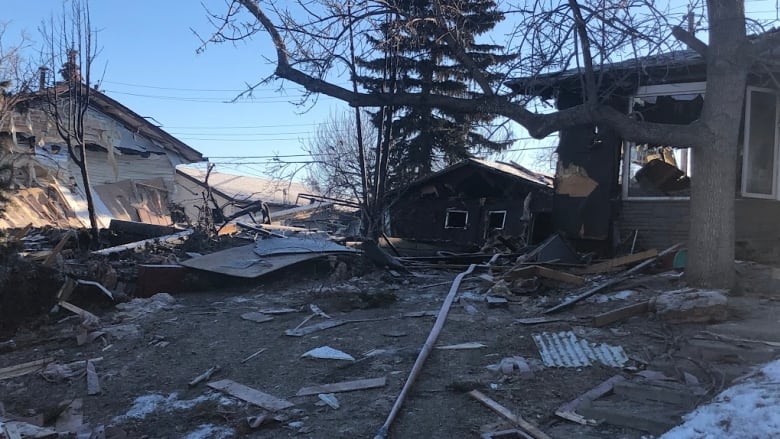 Debris is scatted over a lawn on a space where a house once stood. A house next door bears mark of fire and a blown out window. 