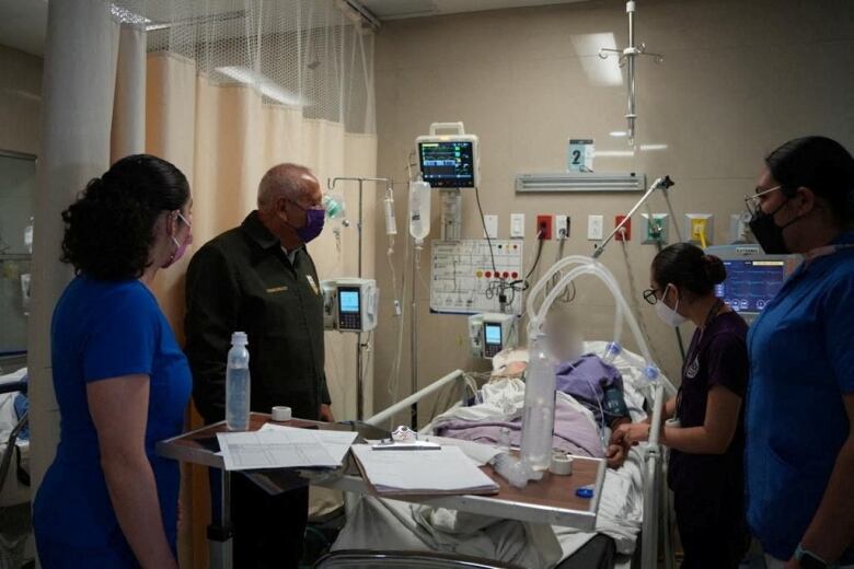 Four people are shown standing near a patient in a hospital room who is lying on a bed.