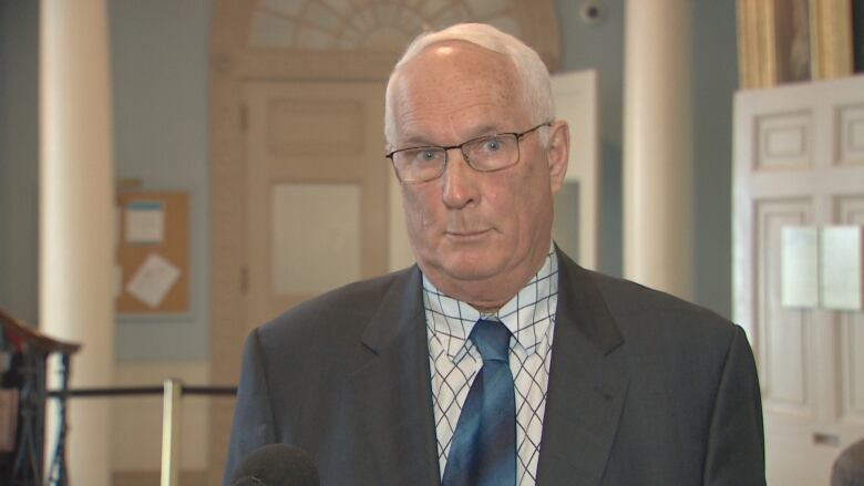 A man with white hair and glasses wears a grey blazer with a white shirt and blue tartan tie.