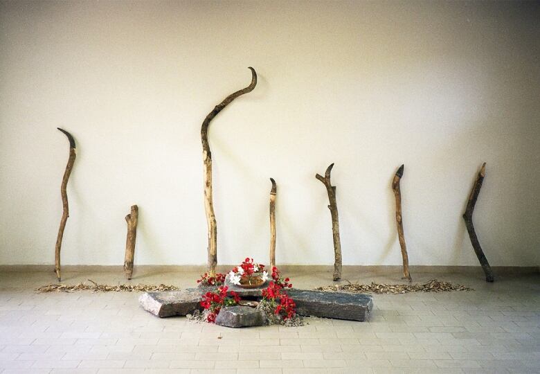 Seven sticks are leaning against a white wall. A line of what appears to be shredded bark is in front of them. In front of that are four slabs of stone in a pile, covered in red and white flowers. On top of the centre stone is a wooden bowl.