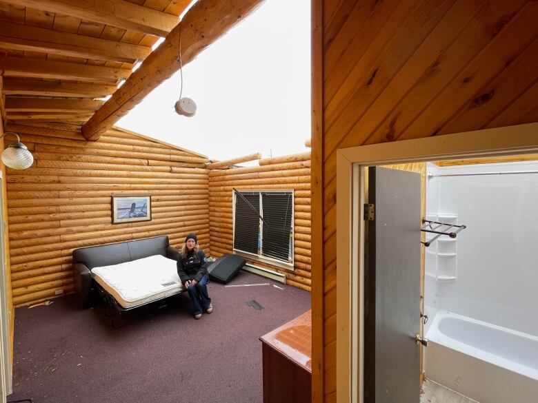 A woman sits on a bed in a cabin that had its roof blown off.