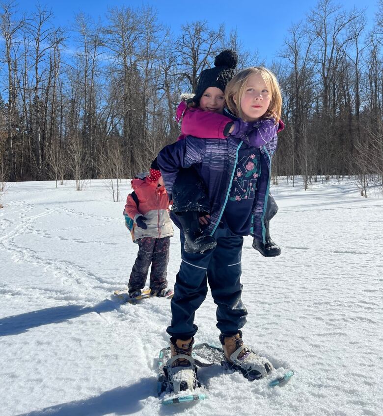 A 9 year old girl wearing snowshoes stands in the snow with a 4 year old girl on her back.