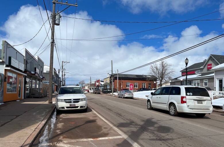 Cars drive down a busy small town Main Street. There is a bank in the background. 