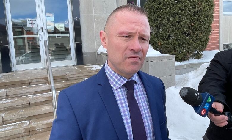 A man with short-cropped hair, wearing a blue suit speaks to reporters outside of the Sudbury courthouse