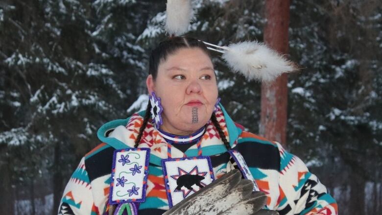 A Cree woman with a chin tattoo is dressed in traditional regalia while standing outside on a snowy day. 