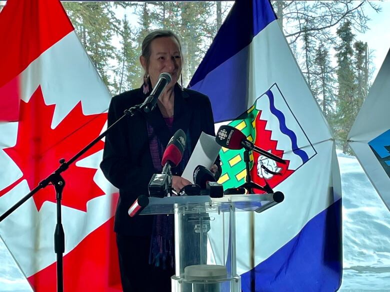 A woman stands smiling at a podium with microphones from the press in front of her and a Canadian flag and Northwest Territories flag behind her.