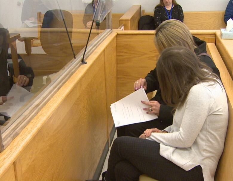 Two women sit on a bench inside a courtroom. One woman holds a document in her hands. 