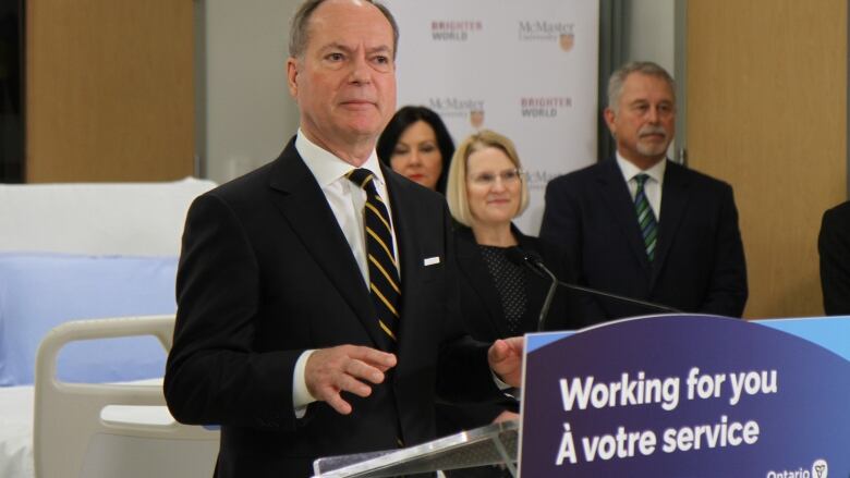 A person in a suit stands behind a podium. Some other people are standing in the background.