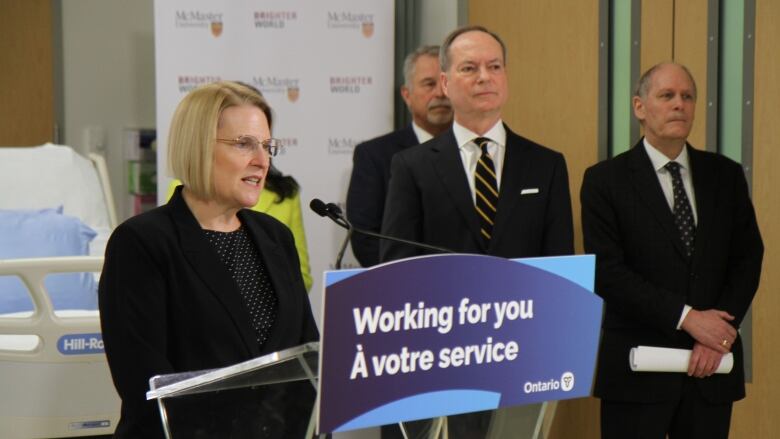 A person in a blazer speaks at a podium, while three people in suits look on.