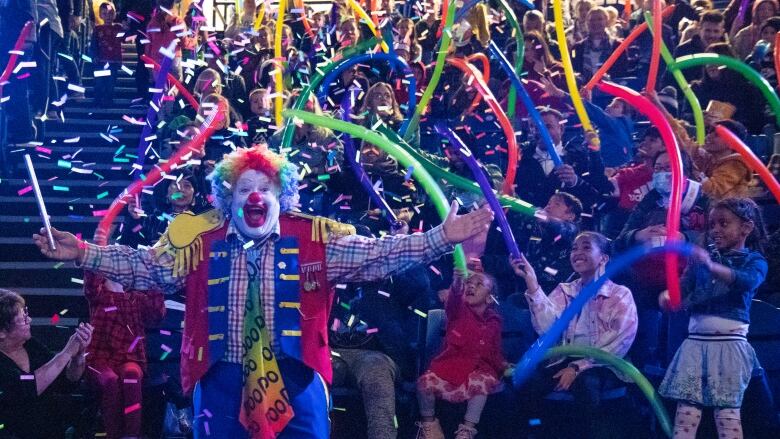 A clown stands in front of audience members holding balloons as confetti rains down.