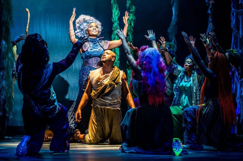 A group of people pose on stage in colourful costumes. In the centre is a man kneeling. Behind him is a smiling woman with upraised arms. 