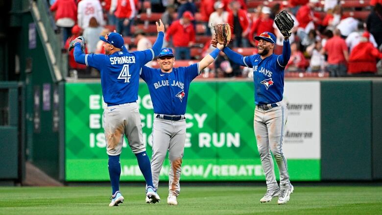 Three male baseball player smile while leaping into the air with their arms raised in a stadium filled with fans.