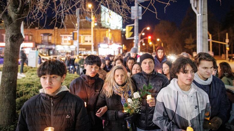 A crowd of people with sad faces walk outside on the sidewalk. Some are holding candles and flowers.