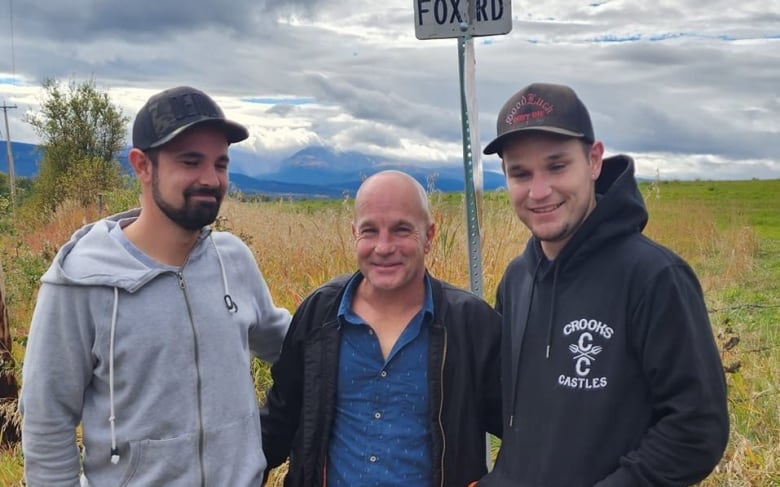 Three men stand together in front of a sign in a field.