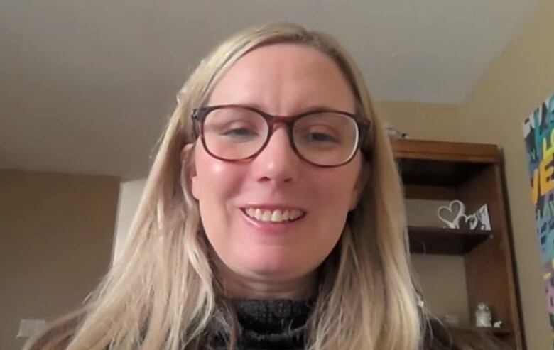 A woman with long hair and glasses smiles at the camera in what appears to be a home office.