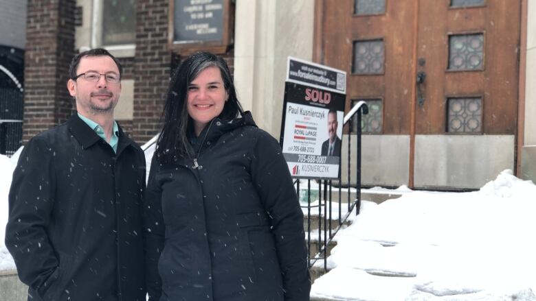 A man wearing a black coat stands beside a woman with dark hair and also wearing a dark coat. They stand in front of a for sale sign at a church.