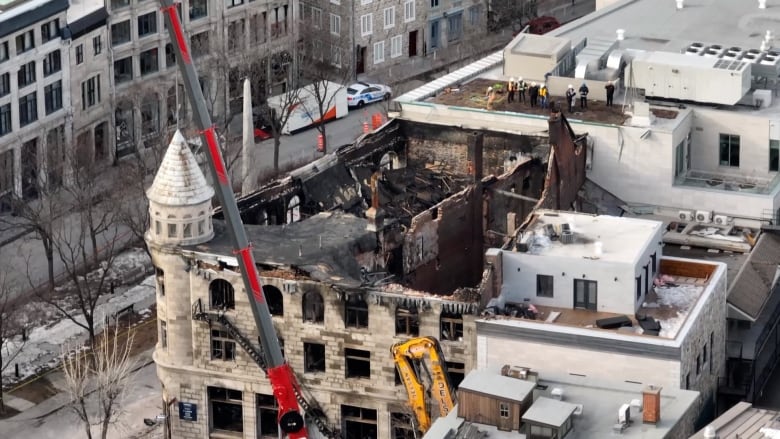 A burned-out building is seen from above, with safety workers and cranes visible.