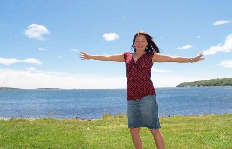 A woman stands on grass with the ocean behind her. She has long dark brown hair and is wearing a red and black top with a denim skirt. Her arms are outstretched and she is smiling.