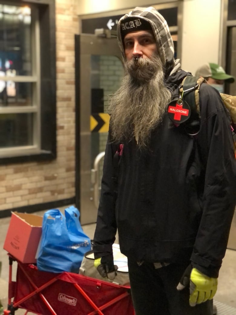 Jay Walls, a tall bearded man, wearing neon gloves, a pin with a red cross that reads naloxone and a hat with the letters ACAB, stands in front of a cart with boxes of supplies. Walls, a volunteer with an independent street team, hands out food and other essentials at Atwater metro on March 22, 2023. 