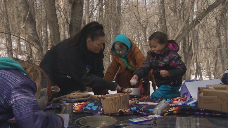 Two children and an adult cook over a fire in the forest.