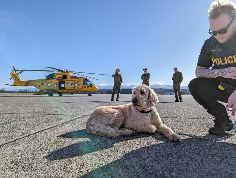 A dog is seen sitting in front of a helicopter while a man kneels beside it.