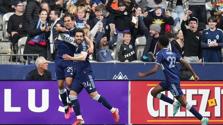 Whitecaps forwards Simon Becher and Brian White embrace in navy blue shirts and shorts, with Whitecaps midfielder Ali Ahmed running towards them. 