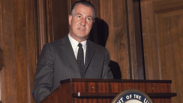 A man in a suit is shown in a panelled room speaking at a podium